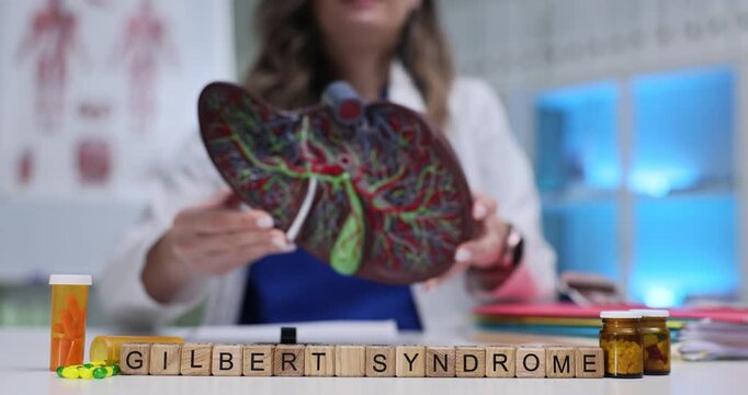 Wooden blocks spell words Gilbert Syndrome near liver model. Woman doctor holds organ model explaining mild jaundice disorder to patient in clinic