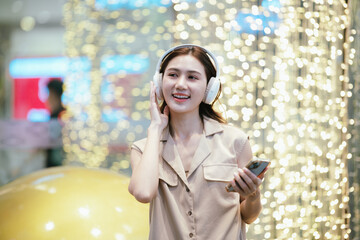A smiling woman enjoying music with headphones while holding her smartphone, surrounded by bright decorative lights in a lively atmosphere.