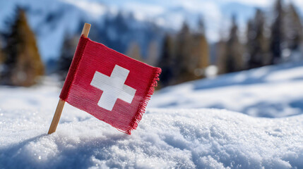 A mini flag of Switzerland in the snow with blurred mountains, a winter travel symbol, a red banner with white cross, a ski resort holiday concept in the Alps.