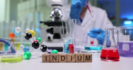 Wooden blocks spell word Indium on bright chemical table in lab. Scientist person in white coat mixes colorful solutions studying metal properties - Powered by Adobe