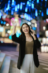 A smiling woman using her smartphone at night in a lively city setting, creating a warm and relaxed atmosphere with colorful blurred lights in the background.