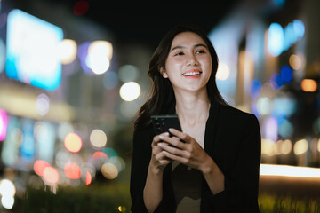 A smiling woman using her smartphone at night in a lively city setting, creating a warm and relaxed atmosphere with colorful blurred lights in the background.