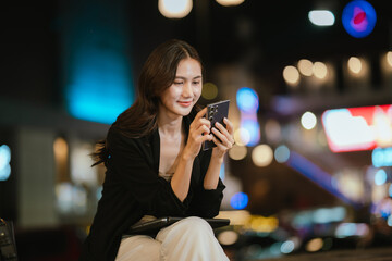 A smiling woman using her smartphone at night in a lively city setting, creating a warm and relaxed atmosphere with colorful blurred lights in the background.