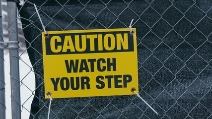 A yellow caution sign reading Caution Watch Your Step mounted on a chain-link fence and secured with white zip ties.