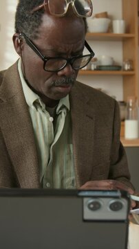 Vertical shot of retired elderly Black man wearing glasses reviewing overdue bills and entering utility amounts into tablet at kitchen table