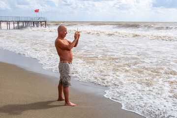 shirtless man standing on sandy beach, taking photo crashing waves with smartphone, barefoot in shallow water, tourist capturing ocean scenery during beach vacation, active retirement lifestyle