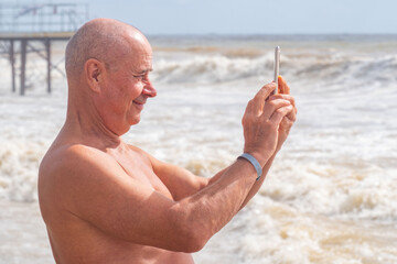 shirtless man standing on sandy beach, taking photo crashing waves with smartphone, barefoot in shallow water, tourist capturing ocean scenery during beach vacation, active retirement lifestyle