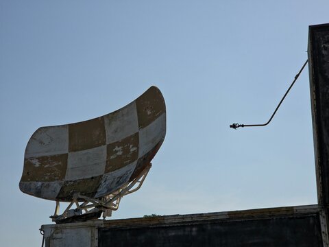 Close-up of a weathered, checkered (white and yellow/brown) radar dish or antenna mounted on a low structure against a clear, light blue sky.