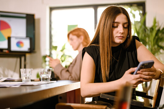 Focused young woman using her smartphone while sitting at a table in a modern office with a colleague working in the background during a busy workday - Powered by Adobe
