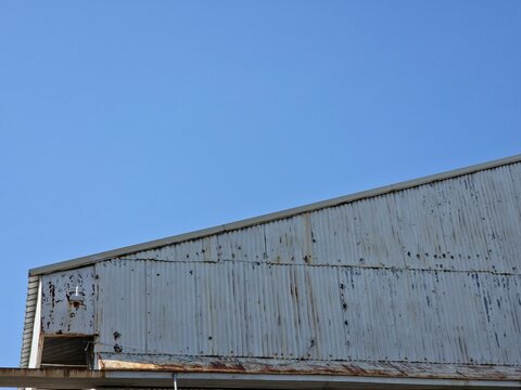 A close-up view of the upper part of a weathered, corrugated metal building or hangar against a clear, bright blue sky. The metal is old, showing signs of rust and peeling white paint.