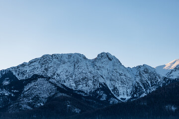 Stunning winter mountain landscape with sunlit snowy summits contrasting against the dark, forested foothills. Clear sky copy space