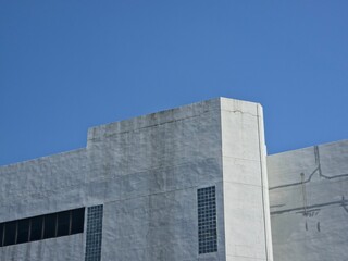 Upper section of a stark, light gray, industrial or commercial building facade with small rectangular windows, set against a clear blue sky.
