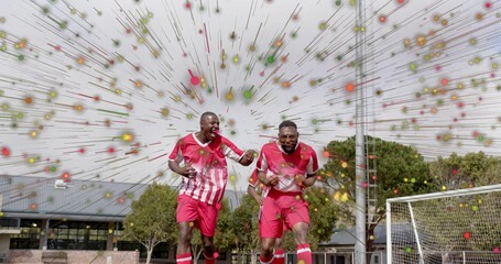 Running two soccer players in red and white uniforms celebrating on grass pitch, with goal net
