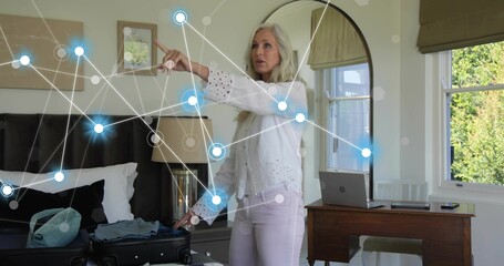 Standing senior woman wearing eyelet top using AR overlay in bedroom with open suitcase and laptop