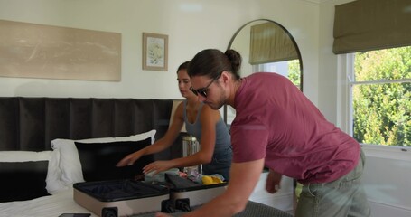 Packing couple wearing maroon t-shirt and gray tank top, loading hard-shell suitcase in bedroom