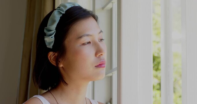 Gazing Korean woman by window at home, wearing pale tank top, pale-green padded headband, necklace
