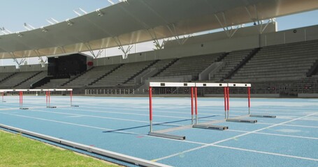 Showing set of red-and-white hurdles crossing blue running track at outdoor stadium, with canopy