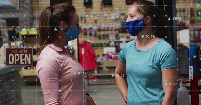 Talking two masked women in light-pink, teal tops standing in small shop with sanitizer, open sign