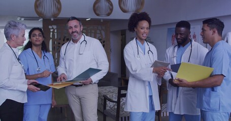 Reviewing charts, six staff in lounge wearing white coats holding folders, tablet, stethoscopes