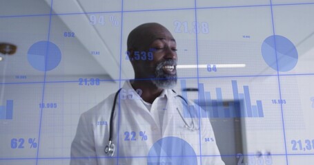 Speaking mature doctor wearing white coat and stethoscope in hospital corridor, blue data overlays