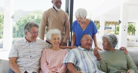Smiling senior group chatting on light-colored sofa near glass sliding doors, wearing sweaters