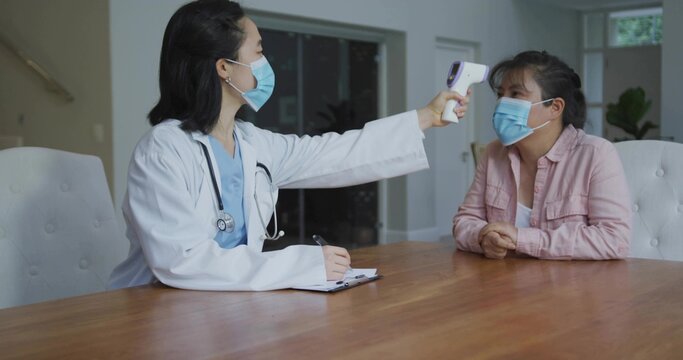 Measuring clinician in white coat, mask checking patient temp at wooden table, with IR thermometer