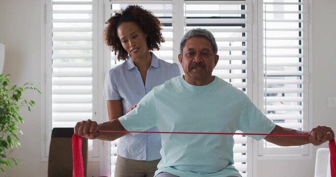 Using red resistance band, senior man wearing mint tee receiving care on chair near shutters