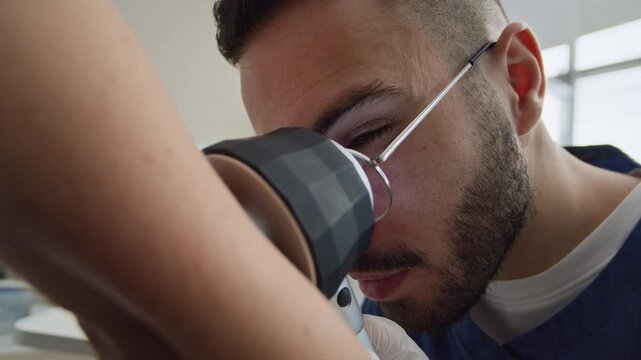 Handheld close up shot of focused young Middle-Eastern male doctor using dermatoscope while looking at skin on unrecognizable patients elbow in clinic