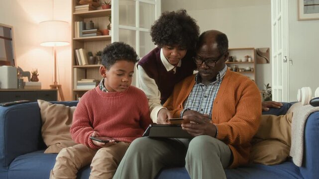 Focused elderly Black man holding credit card learning how to shop online while young grandchildren explaining payment methods to grandfather pointing at tablet screen sitting on couch in living room