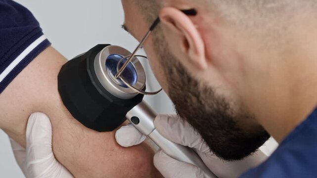 Closeup shot of male doctor in eyeglasses using dermatoscope while examining skin condition on forearm of unrecognizable patient