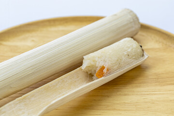 A piece of sticky rice with an orange yam is partially unwrapped from a bamboo casing, resting on a round wooden plate