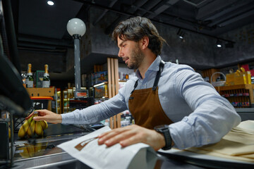 A man in apron diligently works in a local grocery store at the cash register
