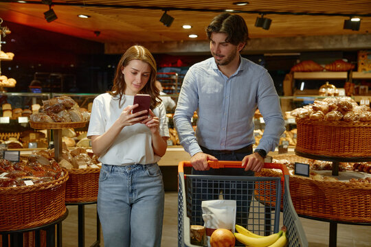 A man is pushing a shopping cart while a woman is looking at her phone