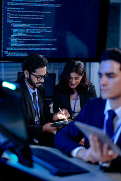 Focused diverse professionals collaborate late night in a modern tech office. Colleagues discuss project details using a tablet with stylus while code runs on a large screen.