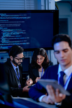 An AI research team brainstorms late night. Innovators discuss machine learning model architecture using a tablet while reviewing training data code displayed on a large screen.
