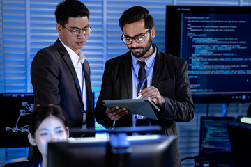 Diverse male professionals strategize late night modern tech hub focused review digital tablet stylus discussing project data code screens intense collaboration synergy achieved.
