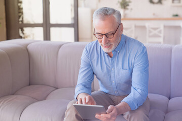 Cheerful elderly grandfather, senior man using digital tablet, searching web, using social media, loading feed, have zoom conference, doing online shopping, calling family at home sitting on the sofa