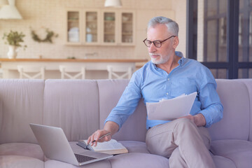 Working serious old senior elderly businessman paying domestic bills online using laptop, counting, paperwork, home office. Social distancing concept. Senior life. Freelance remote work