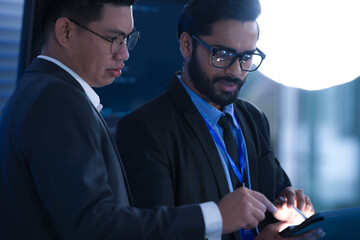 Dynamic tech team collaborates late night. Young Asian woman points at screen explaining project details to male colleague reviewing documents in modern hub workspace.