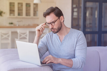 Stressed sad tired serious young caucasian man businessman student professor freelancer working using laptop sitting on the sofa at home indoors. Home office, business problems, remote work