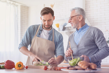 Young adult son preparing meal, food, cutting salad for his old elderly senior father in the kitchen. Cooking food together. I love you, dad! Happy father`s day! Men cooking together