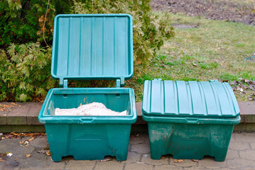 Granular de-icing mixture inside green storage container, winter road treatment material combining sand and salt to enhance traction and prevent slips on icy surfaces. Winter road, pathway maintenance