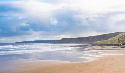 A sandy beach with a rough sea and headlands. A cloudy sky is above.