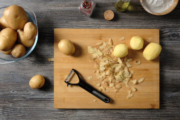 Top view of raw potato tubers in an glass pot, knife for cleaning vegetables, red pepper, flour, butter and spiceson wooden table.