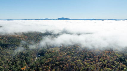Aerial view of fog-covered Adirondack forest with autumn foliage and distant mountain peaks under clear blue sky.