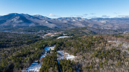 Panoramic winter view of the Adirondack Mountains in New York with forested valleys, snow patches, and clear blue sky.