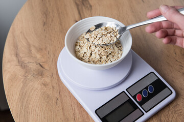Weighing oatmeal in a bowl on a digital kitchen scale, a woman's hand holding a spoon pouring oatmeal into a bowl