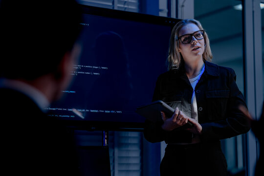 A female software architect presents the system architecture or walks through a complex codebase on a large screen using a tablet to guide her development team during a review.