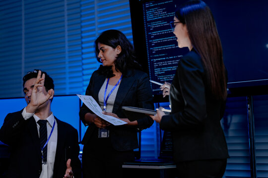 Two female engineers one Asian and one Indian discuss data with a male colleague in a command center. They are analyzing AI code for a cybersecurity system. - Powered by Adobe