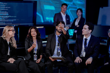 A large IT department meeting in a command center (SOC). An Indian analyst holds a tablet, listening with his team to a cybersecurity update on an AI system.